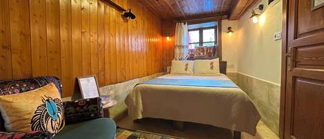 Una habitación acogedora con paredes de madera, una cama con ropa de cama estampada, una mesita con un cuadro enmarcado y un colcha colorida en una silla.