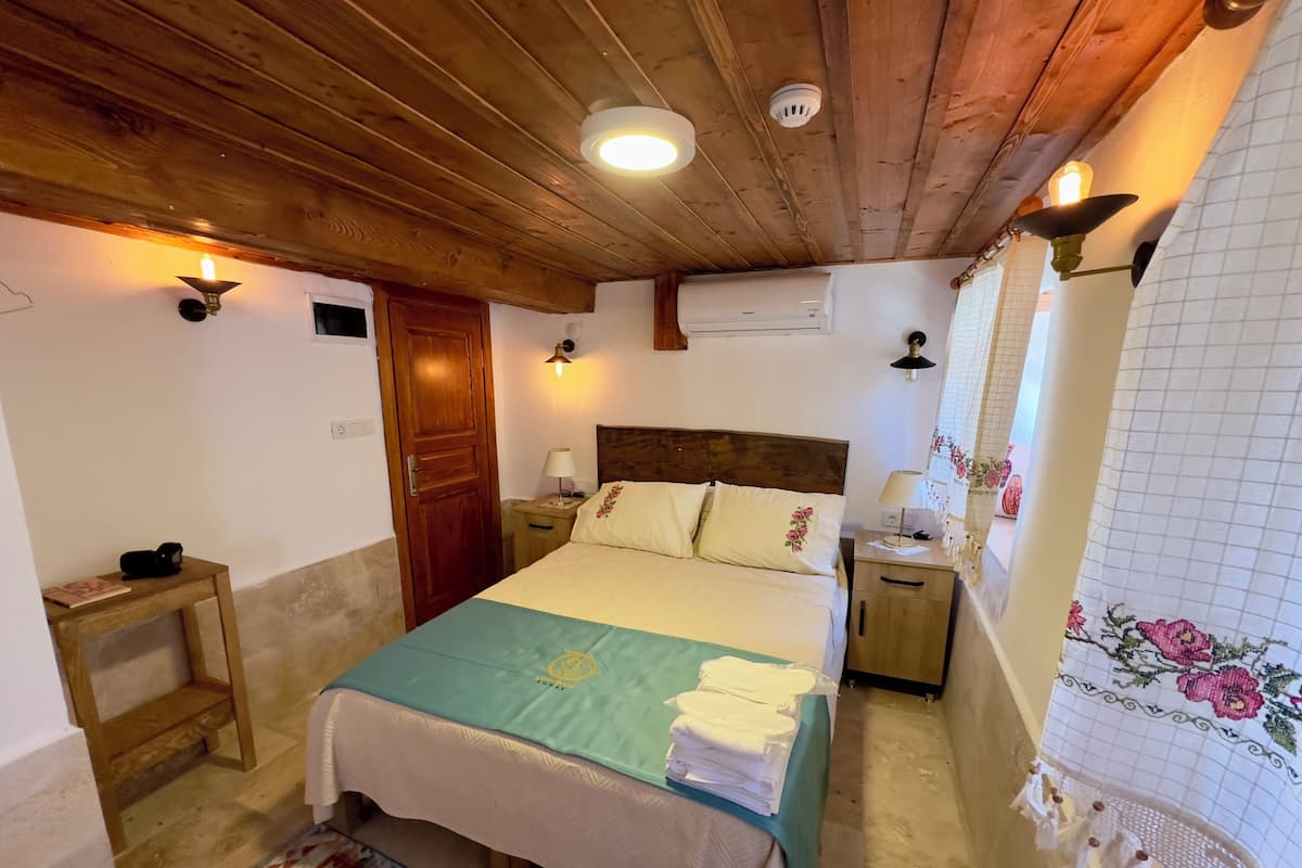 A cozy bedroom with a wooden ceiling, a bed with a green bedspread, a nightstand with a lamp, and a window with a floral curtain.