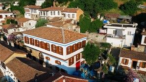 A two-story building with a red-tiled roof, white walls, and wooden shutters. It has a balcony with blue chairs and a small garden area. - Çirkince Konak Şirince (Selçuk)
