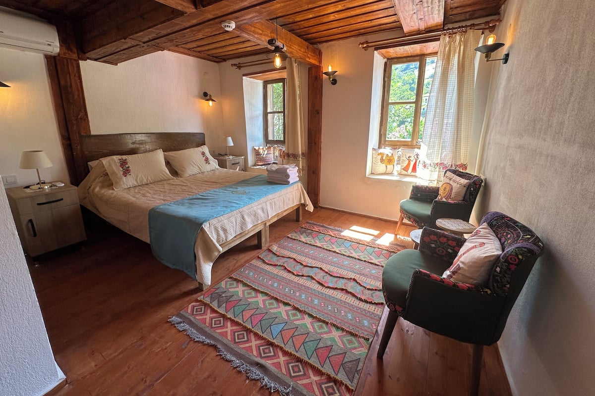 A cozy bedroom with a wooden ceiling, a large bed with a blue bedspread, a nightstand with a lamp, a chair with colorful cushions, and a window with curtains.