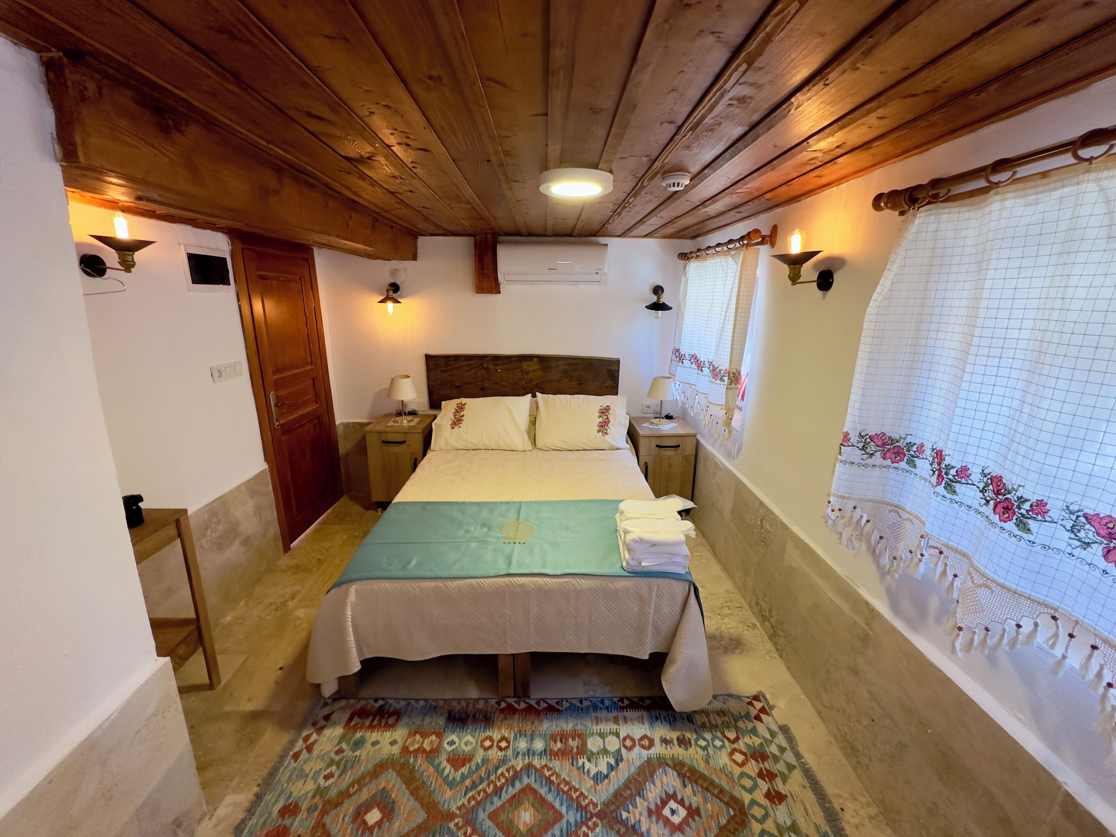 A bedroom with a wooden ceiling, a bed with a patterned bedspread, two bedside tables with lamps, a patterned rug, and a window with a floral curtain.