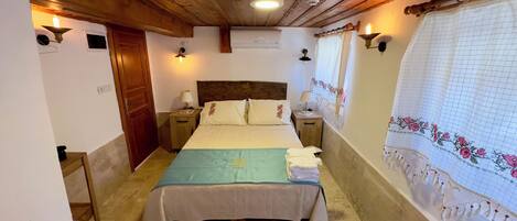 A bedroom with a wooden ceiling, a bed with a patterned bedspread, two bedside tables with lamps, a patterned rug, and a window with a floral curtain.