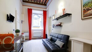 A cozy living room with a black leather sofa, a small white table, a TV mounted on the wall, and a window with red curtains. - Mansion in Lucca Near Via Fillungo (Lucca)