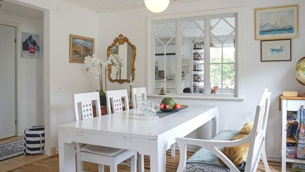 A dining area with a white table, chairs, and a window with a view of a house and trees. - 4 Star Holiday Home in Brastad (Brastad)