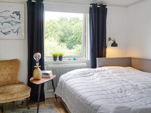 A bedroom with a bed, a chair, a small table with a lamp, a radiator, a window with curtains, and a wall-mounted picture of whales. - 4 Star Holiday Home in Brastad (Brastad)