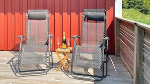 Two transparent chairs with metal frames, a bottle of champagne, and two glasses on a wooden deck against a red wall, with a Swedish flag in the background. - 6 Person Holiday Home in Mardaklev-by Traum (Mårdaklev)