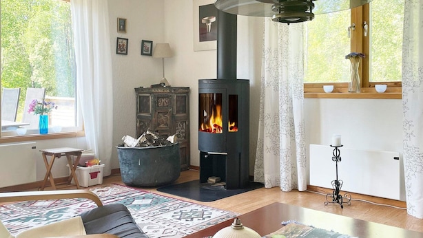 A cozy living room with a fireplace, a dining table with a vase and remote controls, and a window with a view of trees. - 6 Person Holiday Home in Mardaklev-by Traum (Mårdaklev)