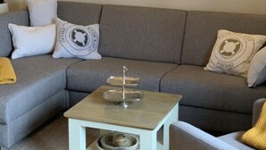 A living room with a grey sofa, a small white side table, and a painting of sailboats on the wall. - Holiday Apartment Harbor Flair (Karlshagen)