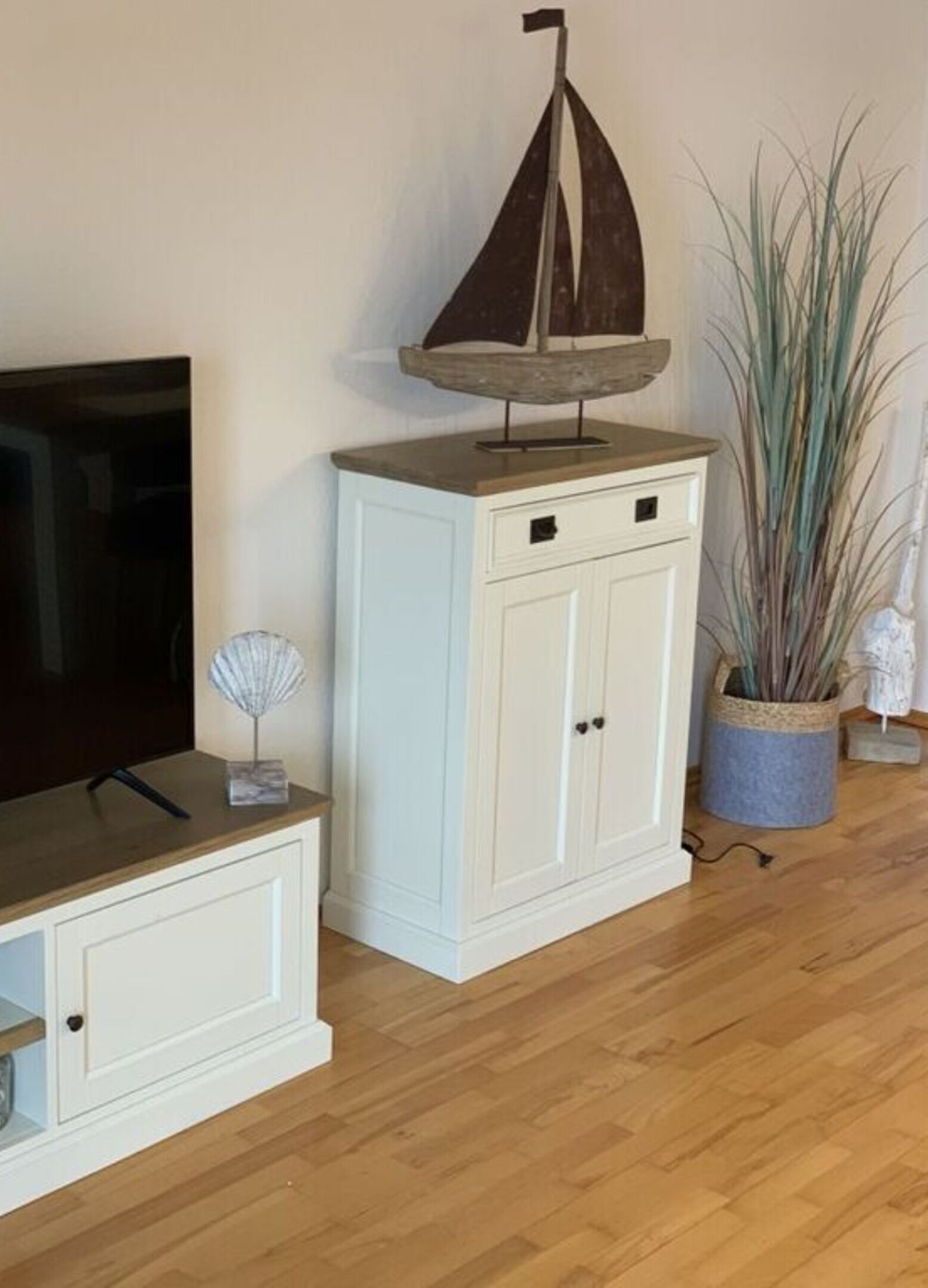 Un meuble blanc à deux portes et une étagère, un téléviseur posé sur une étagère inférieure, un petit voilier décoratif et une grande plante dans le coin.
