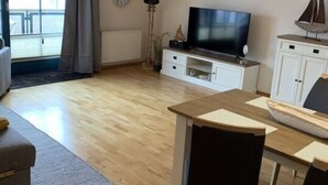 A modern living room with a wooden floor, a sofa, a dining table with chairs, a TV, and a window with curtains. - Holiday Apartment Harbor Flair (Karlshagen)