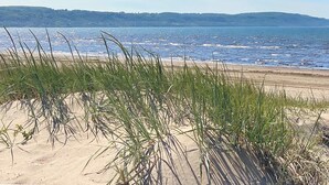 Una playa con dunas de arena y pasto en primer plano, que se extiende hasta un mar tranquilo y colinas a lo lejos.