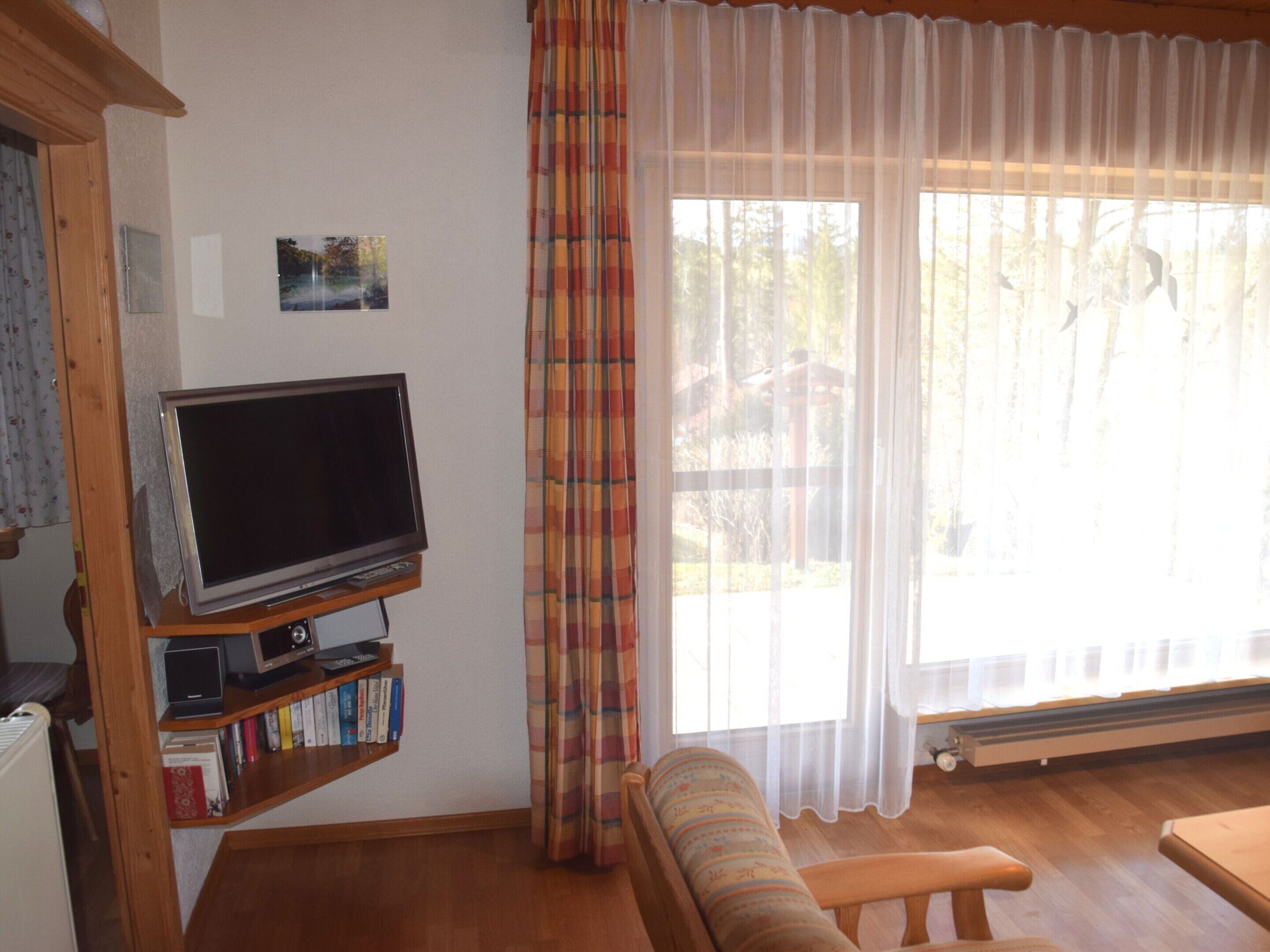 A living room with a television on a wooden stand, a bench, and a window with curtains.