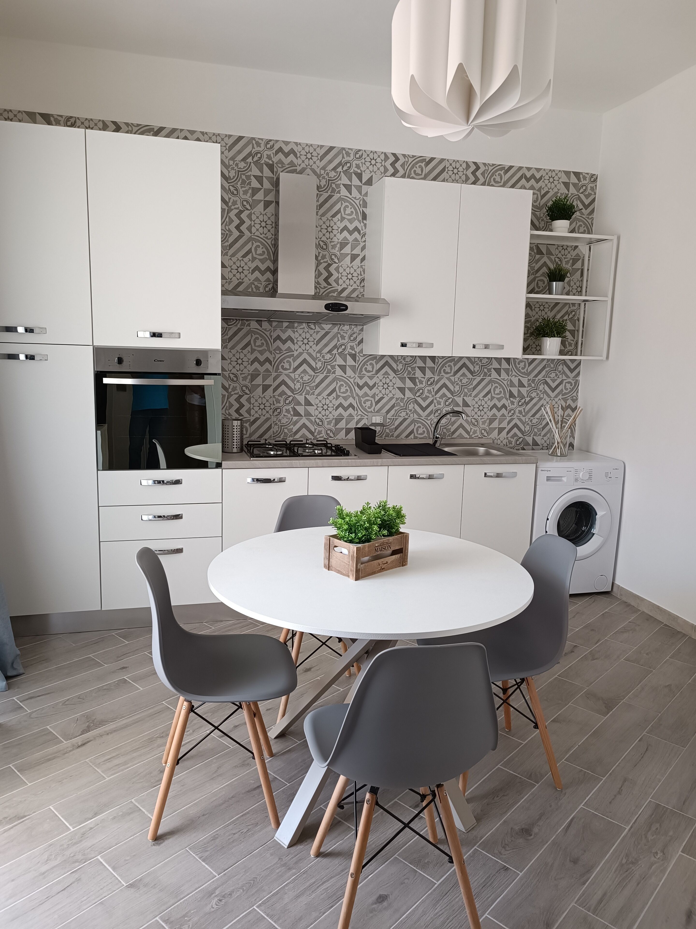A modern kitchen with white cabinets, a built-in oven, a gas stove, a sink, and a washing machine. There is a round table with four chairs and a potted plant on it.