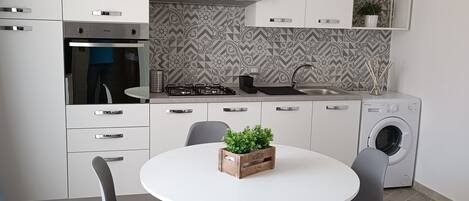 A modern kitchen with white cabinets, a built-in oven, a gas stove, a sink, and a washing machine. There is a round table with four chairs and a potted plant on it.
