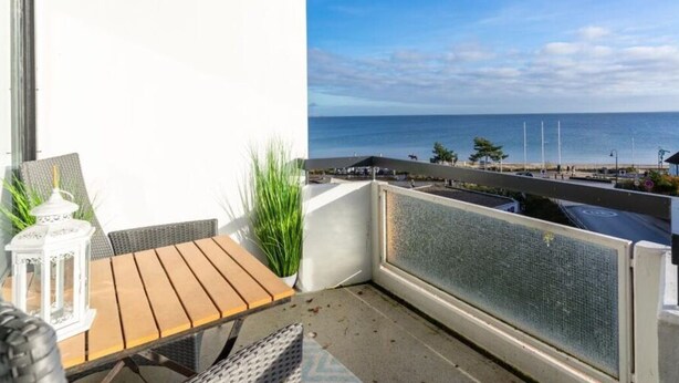 Ein Balkon mit Holztisch und -stühlen, einer weißen Laterne und Blick auf Meer und Strand.
