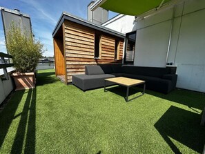 A modern outdoor patio with a wooden deck, a grey sofa, a small table, and a potted plant. - 2-room Luxury Apartment (Scharbeutz)