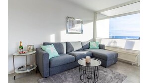 Ein modernes Wohnzimmer mit einem grauen Ecksofa, einem kleinen runden Couchtisch und einem Beistelltisch mit einer Flasche und einem Glas.