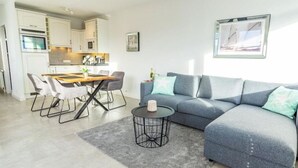 Ein modernes Wohnzimmer mit einem grauen Sofa, einem Esstisch mit Stühlen, einer Küchenzeile mit weißen Schränken und einem gerahmten Bild an der Wand.