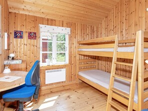 A room with wooden walls, a bunk bed, a desk with a chair, a window with a view of greenery, and a radiator.