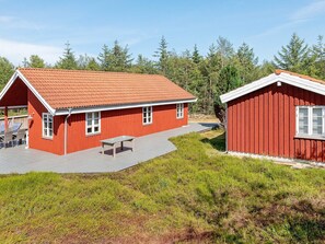 Une cabane en bois rouge avec un toit en tuiles, une terrasse avec une table et des chaises, et un petit abri de jardin.