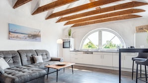 A modern living space with a grey sofa, a wooden coffee table, and a kitchen area with white cabinets and a large window. - No. 7 Lake View Modern Retreat (Aurich)