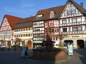 En traditionel europæisk byplads med bindingsværkshuse, en springvand og et skilt for 'Stern - Apotheke'.