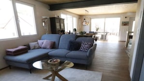 Un salon moderne avec un canapé d’angle, une table basse en verre et un coin repas à l’arrière-plan.