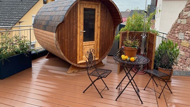 Une structure en forme de tonneau, faite de bois, avec une porte, une petite table entourée de chaises et des plantes en pot disposées sur une terrasse en bois.