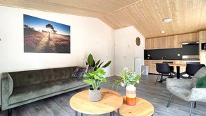 A modern living room with a sofa, two round tables with plants, a kitchen in the background, and a large landscape painting on the wall. - Chalet w/ Garden & Views (Hoenderloo)