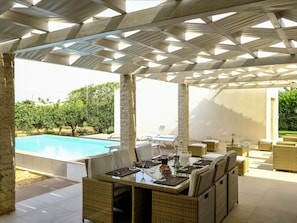 Een terras buiten met een zwembad, rieten meubels en een pergola.