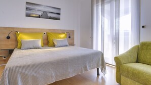 A modern bedroom with a wooden headboard, a large bed with striped bedding, two yellow pillows, a green armchair, and a cityscape painting on the wall. - Nicely Decorated Apartment With Private Pool (Vodnjan)