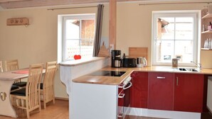 A compact kitchen with a dining area, wooden chairs, a small island with a sink, and a window with a view of a wooden deck. - Holiday Apartment Schiechtele 2 (Pfronten)
