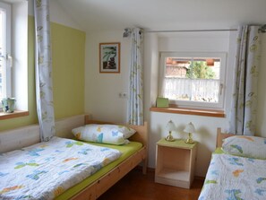 A room with two wooden beds, a small bedside table with lamps, a window with a view of greenery, and a picture on the wall. - Holiday Apartment Schiechtele 2 (Pfronten)