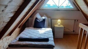 A small, cozy attic bedroom with a sloped ceiling, a single bed, a nightstand with a lamp, and a view through a skylight. - Luetje Huuske Modern Retreat (Krummhoern)