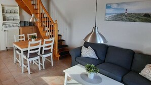 A living room with a wooden staircase, a dining area with white chairs, a grey sofa, and a painting of a lighthouse on the wall. - Luetje Huuske Modern Retreat (Krummhoern)