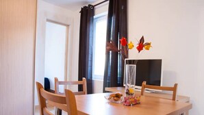 Un comedor con una mesa de madera, cuatro sillas, un florero con flores, un plato de comida y un televisor.