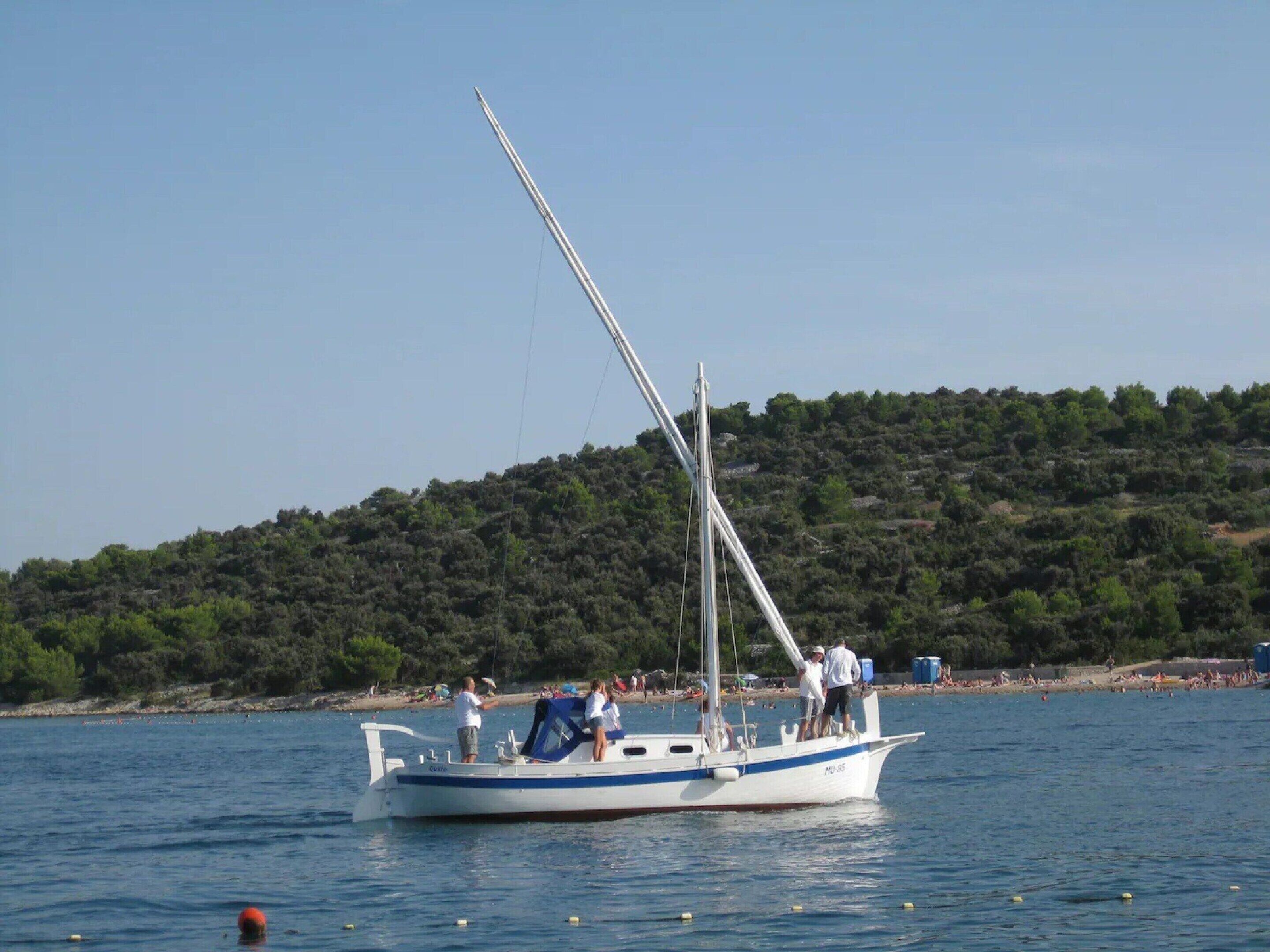 A sailboat with people on board, docked near a beach with a forested hill in the background.