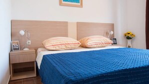 Una cama bien hecha con un edredón azul, dos almohadas a rayas, un cuadro enmarcado con una escena costera, una lámpara de noche, un pequeño estante con un objeto enmarcado, y un jarrón con flores amarillas.