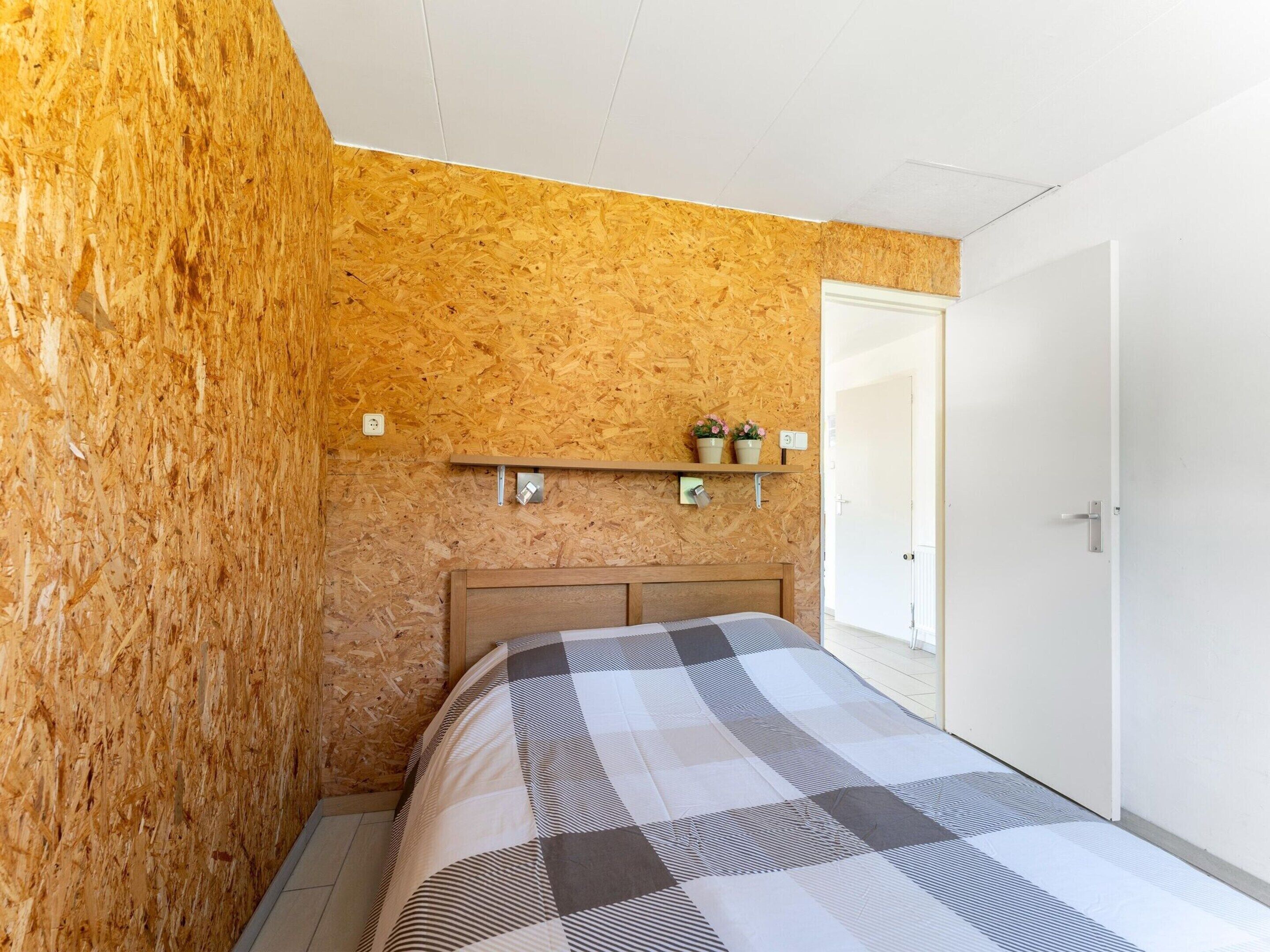 A bedroom with a wooden wall, a bed with a checkered bedspread, a shelf with two potted plants, and a white door.