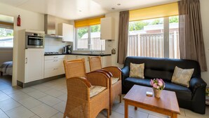 A compact living space with a kitchenette, a dining area with wicker chairs, a black sofa, and a wooden table. - Holiday Home in Baarland With Garden (Baarland)
