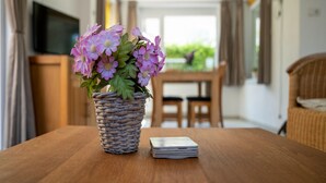 A wicker basket with purple flowers on a wooden table, a stack of cards, and a television in the background. - Holiday Home in Baarland With Garden (Baarland)