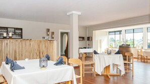 Uma sala de jantar ampla, com piso de madeira, toalhas de mesa brancas e cadeiras de madeira simples.