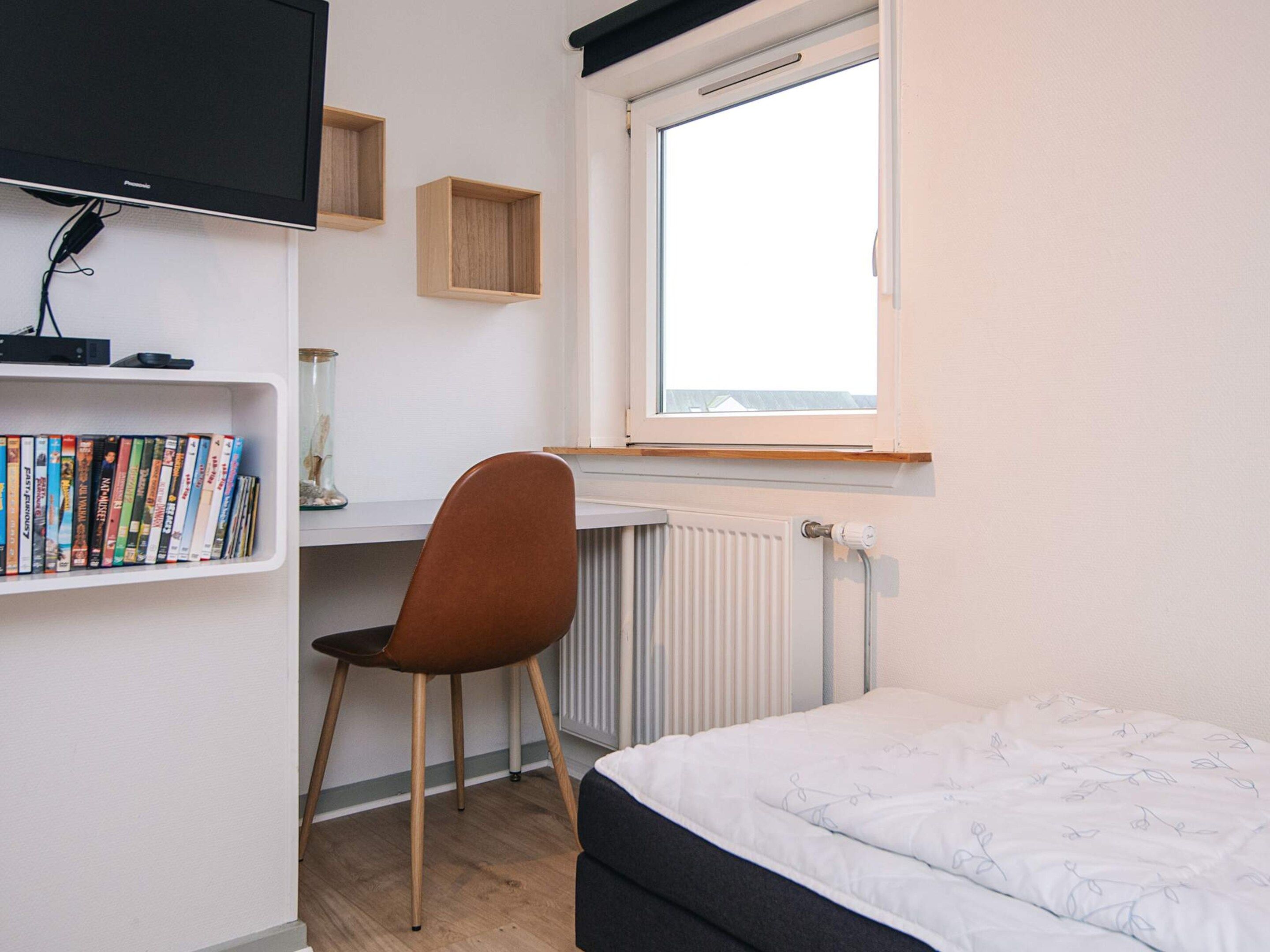 A bedroom with a bed, a desk with a chair, a TV mounted on the wall, and a window.