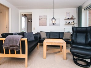 Uma sala de estar moderna com um sofá de couro preto, uma mesa de centro de madeira e uma poltrona de couro preto. Há duas obras de arte emolduradas na parede, uma luminária pendente e uma pequena prateleira com objetos decorativos.