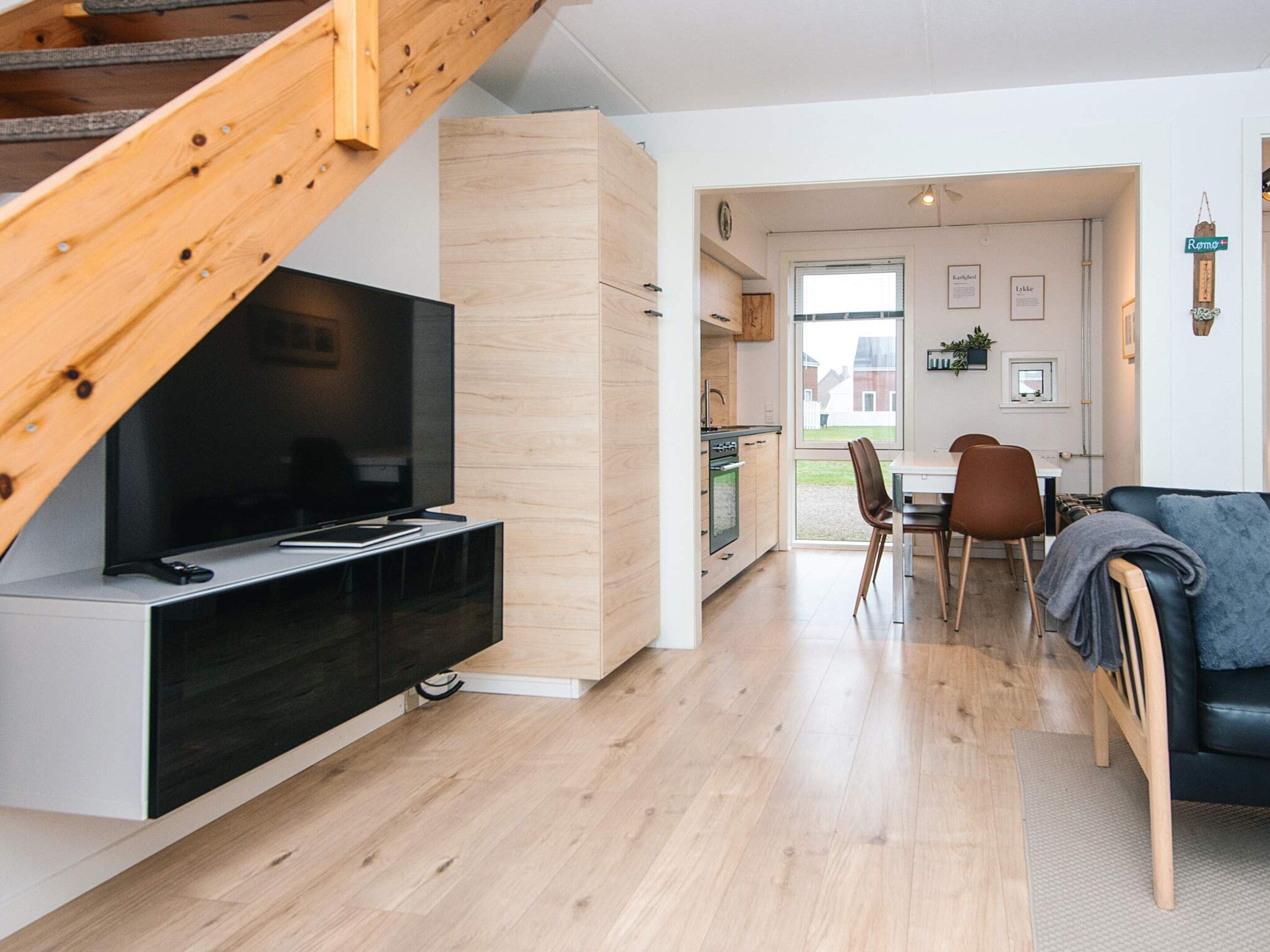 A modern living space with a TV on a black stand, wooden staircase, a dining area with chairs, and a kitchen visible through an open door.