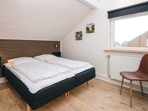 A modern bedroom with a sloped ceiling, a bed with white bedding, a wooden headboard, a black bench, a brown chair, and two paintings on the wall.