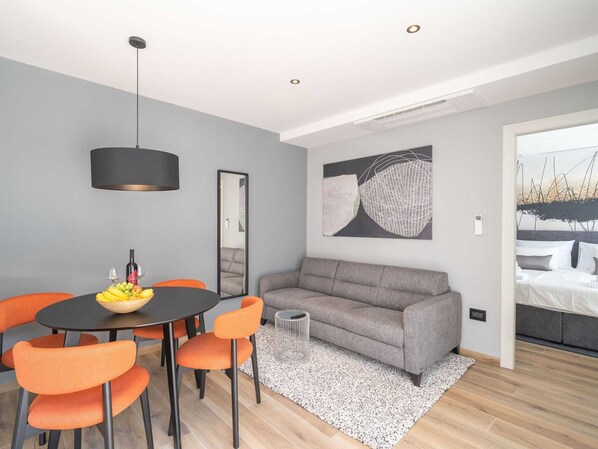 A modern living room with a grey sofa, orange chairs, a round dining table with a fruit bowl, and a large abstract painting. - Apartments Villa Royale - Luxury One Bedroom Apartment With Terrace (Konavle)