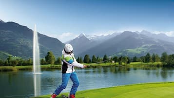 Eine Person fährt auf einer grünen Piste, im Hintergrund sind Berge und ein See zu sehen.