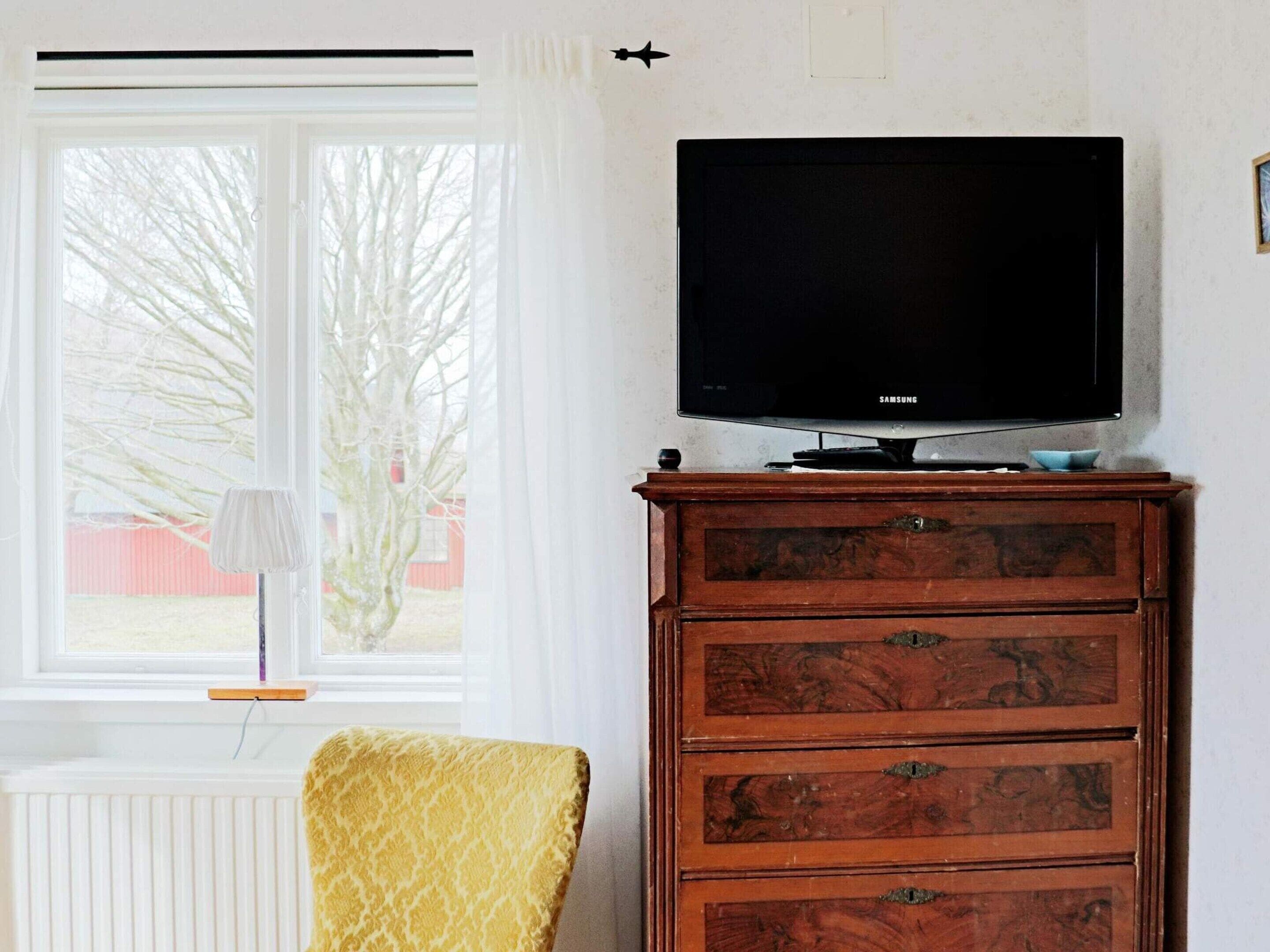 Una habitación con una televisión sobre un mueble de madera, una ventana con vistas a árboles y un radiador.