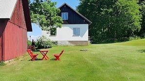 A house with a white exterior and dark roof, a red barn to the left, and a grassy lawn with red chairs and a table. - 9 Person Holiday Home in Bastad (Bastad)